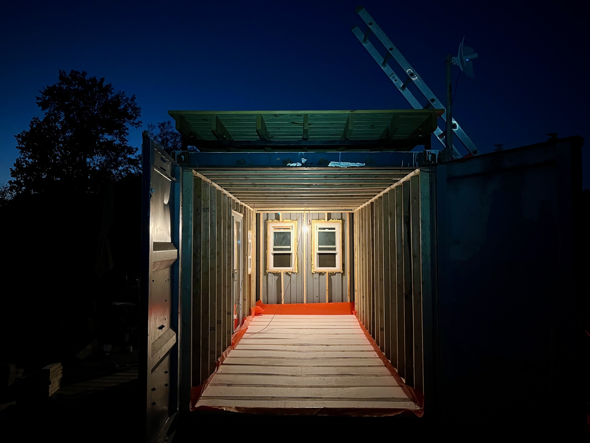 A small, illuminated shipping container cabin under deep twilight, its framed walls glowing warm against the night.