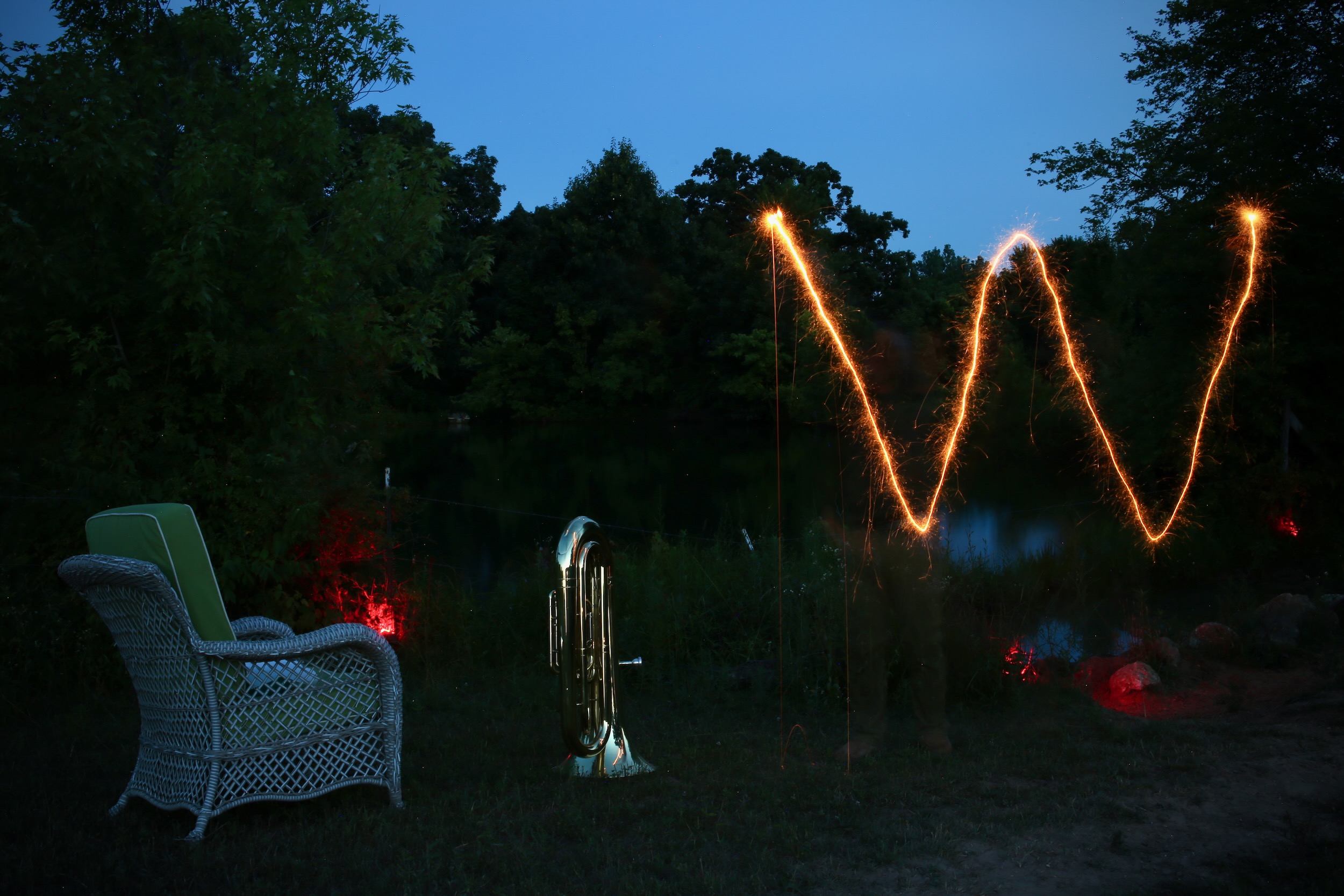 A sparkler burning in a dark field, its sparks mirrored by distant fireflies. A tuba rests nearby in moonlight.